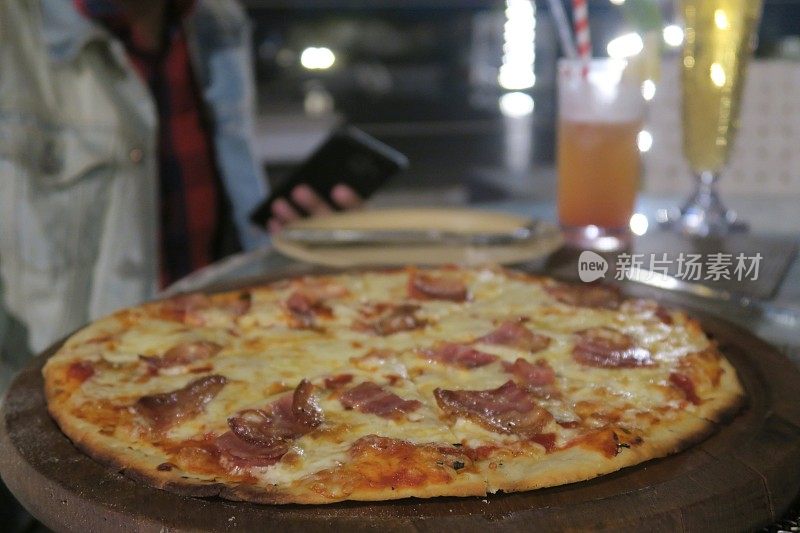 薄而脆的意式烟肉披萨配上金色融化的莫扎雷拉奶酪，在意大利餐厅露天用餐，在酒吧花园，晚上饥饿的用餐者拿着手机喝着啤酒/鸡尾酒，吃着美味的披萨